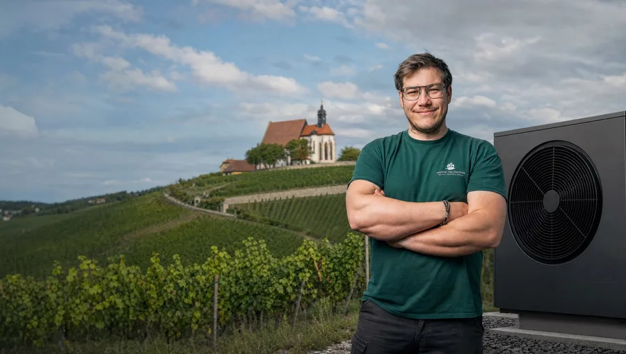 Nico Wehner neben einer installierten Wärmepumpe, dahinter Weinberge